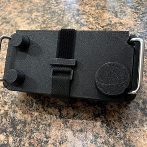May include: A black plastic box with two round knobs, a black strap, and two silver metal handles. The box has a textured surface.