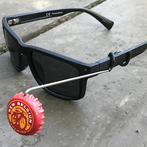 May include: Black sunglasses with a metal hook attached to the arm. The hook is holding a red bottle cap with the words "New Belgium Brewing" and a bicycle logo.