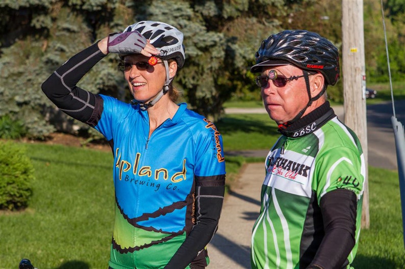 May include: Two people wearing cycling helmets and jerseys are standing on a sidewalk. The person on the left is wearing a blue and green jersey with the text "Upland Brewing Co." The person on the right is wearing a green and white jersey with the text "Blackhawks Bike Club".