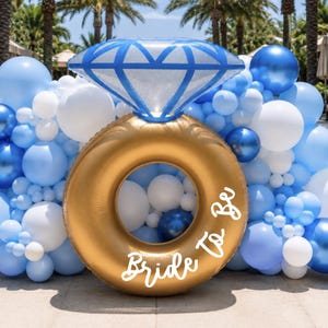 May include: A large inflatable gold ring balloon with a blue diamond-shaped topper, featuring the white text "Bride To Be" on the front. The ring is surrounded by a dense cluster of various sized balloons in shades of light blue, white, and dark blue.