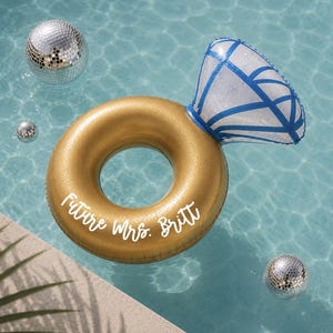 May include: An inflatable pool float shaped like a gold engagement ring with a large diamond topper featuring blue geometric lines. The gold band has the white text Future Mrs. Britt printed on it. Three small silver disco balls float nearby.