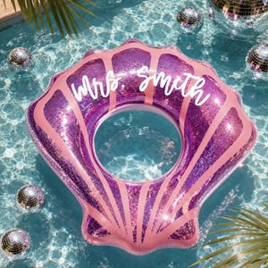 May include: A purple glittery seashell-shaped pool float with pink accents and the text "Mrs. Smith" written in white script across the top. The inflatable pool accessory features a central circular opening and a scalloped shell design.