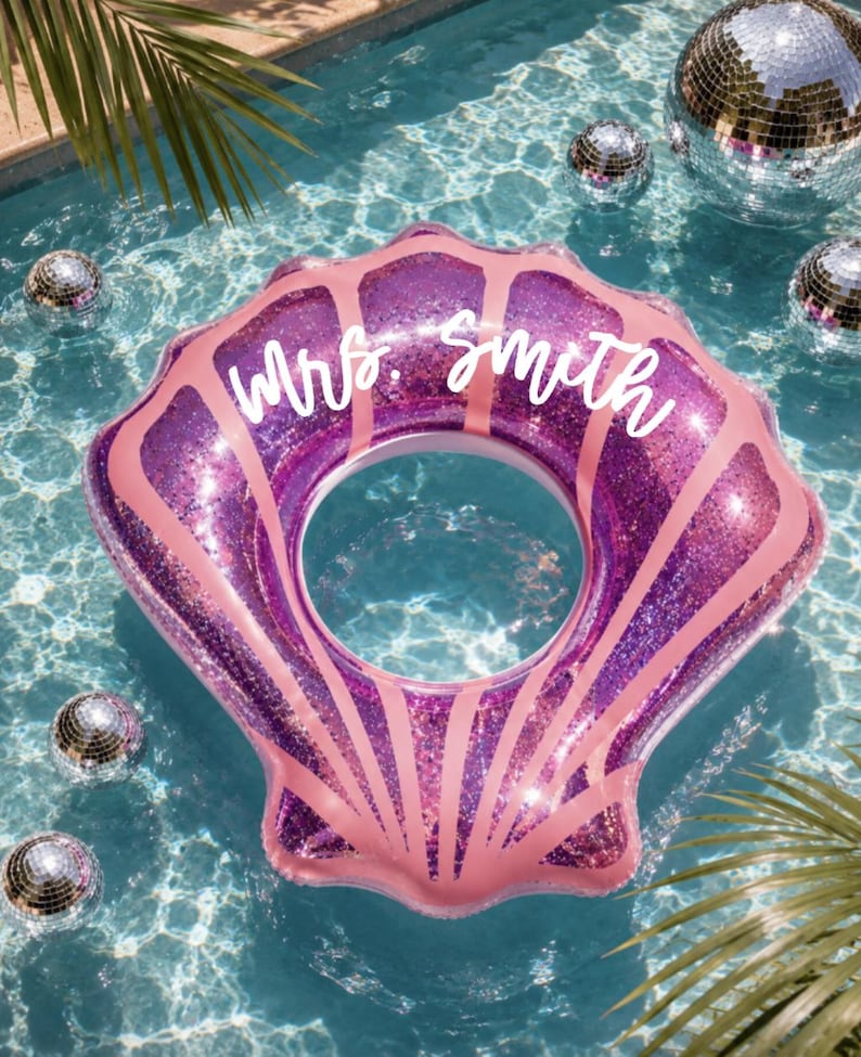 May include: A purple glittery seashell-shaped pool float with pink accents and the text "Mrs. Smith" written in white script across the top. The inflatable pool accessory features a central circular opening and a scalloped shell design.