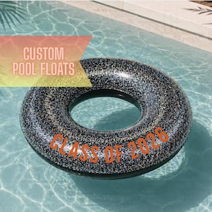 May include: A black and silver glitter pool float with the text "CLASS OF 2026" in orange. The words "CUSTOM POOL FLOATS" are in the background. The float is in a pool with clear blue water.