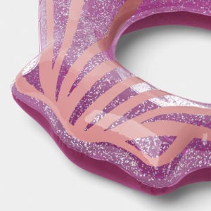 May include: An inflatable pool float shaped like a seashell, featuring a translucent purple material embedded with silver glitter. The surface is decorated with light pink, curved, radiating stripes that mimic the ridges of a scallop shell.