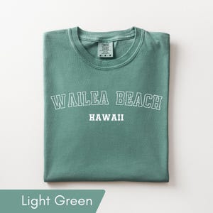May include: A light green short-sleeve t-shirt featuring the white text WAILEA BEACH in a collegiate font arched over the word HAWAII. The shirt has a crew neckline and a Comfort Colors brand label printed on the inside of the collar area.