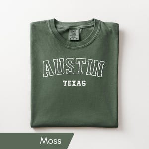 May include: A moss green short-sleeved t-shirt featuring the white text AUSTIN in a large arched collegiate font above the word TEXAS in a smaller block font. The shirt has a crew neckline and a Comfort Colors brand label at the inner neck.