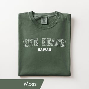 May include: A moss green short-sleeve t-shirt featuring the white text KE'E BEACH HAWAII printed in a collegiate font across the chest. The shirt has a crew neckline and a Comfort Colors brand label visible on the inside of the collar.