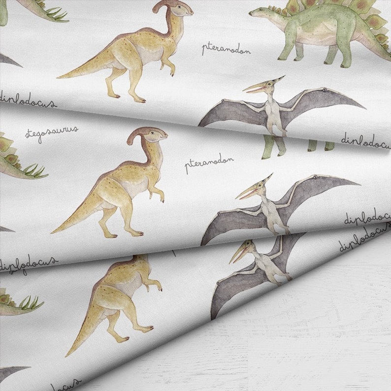 May include: White fabric with a repeating pattern of watercolor dinosaurs. The design includes various dinosaurs such as the Parasaurolophus, Stegosaurus, and Pteranodon, with their names written in cursive.