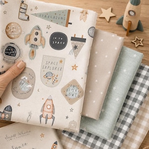 Puede incluir: Una pila de telas dobladas que incluye una pieza de color crema con ilustraciones temáticas del espacio, como cohetes, planetas, estrellas y el texto "STARGAZING," "IN TO SPACE," "SPACE EXPLORER," y "OUT OF THIS WORLD," junto a telas lisas y a cuadros.
