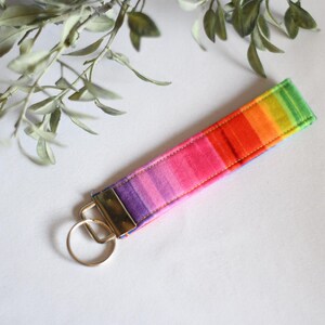 May include: A rainbow-colored wristlet keychain with a gold-tone metal clasp and key ring. The wristlet features a vibrant gradient of colors, transitioning from green to red and purple. The accessory is set against a white background.