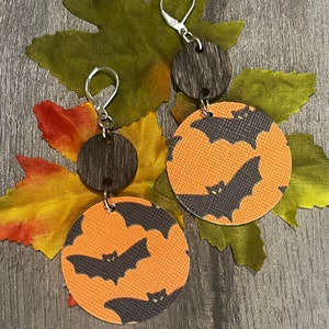 May include: Halloween-themed earrings featuring a dark wood disc and a larger orange circle with a repeating black bat pattern. The earrings have silver-colored ear wires and are displayed on a leaf.