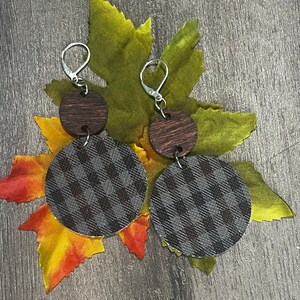 Può includere: Orecchini pendenti con ganci color argento. Ogni orecchino presenta un piccolo cerchio di legno marrone scuro sopra un pendente circolare più grande con un motivo a quadri grigio e nero. Gli orecchini poggiano su foglie autunnali.