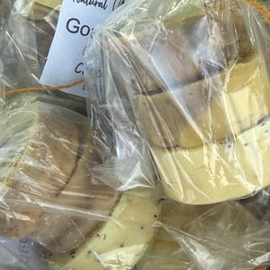 May include: Close-up of several bars of natural goat milk soap, individually wrapped in clear plastic. The soap is a light yellow color with dark specks. A tag reads "Natural Goat". The soap is tied with twine.