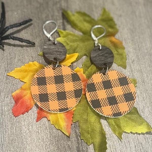 Può includere: Orecchini pendenti a tema Halloween. Gli orecchini presentano una parte superiore in legno scuro e un motivo a quadri rotondi arancione e nero. Gli orecchini sono esposti su foglie autunnali con un ragno di plastica.