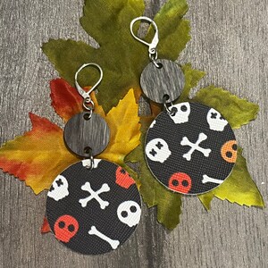May include: A pair of Halloween-themed earrings. Each earring features a small, round, gray wooden top piece and a larger, circular bottom piece with a black background. The bottom piece is decorated with white skulls, orange skulls, and white crossbones.