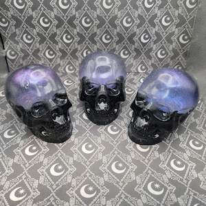 May include: Three black resin skull figurines with a galaxy-like purple and blue glitter embedded in the resin.