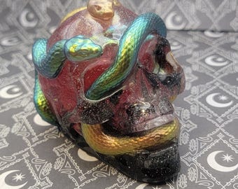 Puede incluir: Una escultura de calavera de resina con una serpiente iridiscente verde y dorada envuelta a su alrededor. La calavera es de color rojo intenso con purpurina negra y plateada incrustada en la resina.