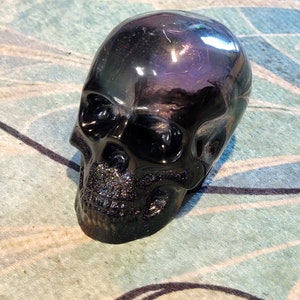 May include: A black skull figurine with a glittery finish. The skull is made of a smooth, shiny material and has a dark, iridescent sheen.