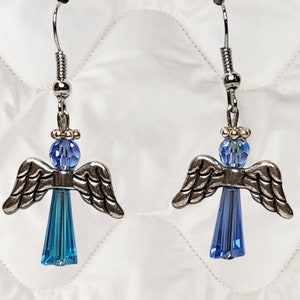 May include: A pair of angel earrings with silver-toned wings and blue crystal bodies. The earrings have a silver hook and a small blue bead at the top of the angel's head. The background is a white, quilted fabric.