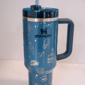 Puede incluir: Vaso Stanley azul con asa y pajita. El vaso presenta un diseño plateado con varias ilustraciones y el logotipo de Stanley. El vaso está diseñado para mantener las bebidas frías o calientes. El vaso mide aproximadamente 25 cm de alto.