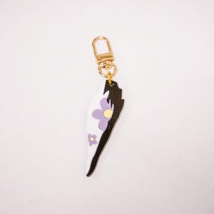 May include: A decorative keychain with a gold-colored clasp. The charm is shaped like a stylized wing, featuring a black and white design with purple flowers and yellow centers. The keychain is a unique accessory.
