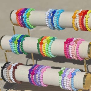 May include: A display of colorful beaded bracelets, each featuring a cross charm. The bracelets are arranged on a gold-toned tiered stand. Colors include red, orange, yellow, green, blue, purple, pink, and brown.