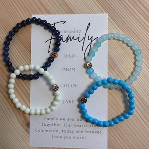 Puede incluir: Cuatro pulseras de cuentas en azul marino, blanco, azul claro y turquesa, con abalorios iniciales de color cobre. Se ve una tarjeta con la palabra "Family" y nombres. La tarjeta también incluye el texto "Family we are, away together."