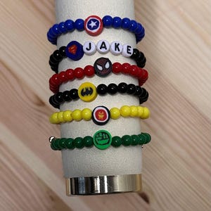 May include: A collection of beaded bracelets in various colors, each featuring a superhero emblem. One bracelet spells out "JAKE" with letter beads. The bracelets are displayed on a white cylindrical stand.