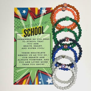 May include: A collection of beaded bracelets in various colors, including orange, green, blue, and white, each adorned with a superhero-themed charm. A colorful card with the text "FIRST DAY OF SCHOOL" and a motivational message is also present.