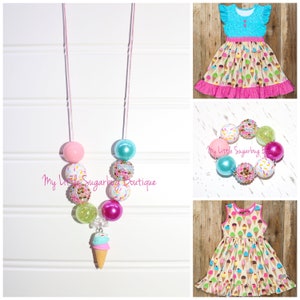 May include: A set of ice cream themed jewelry for girls, including a necklace with a pink, green, blue, and white bead design and an ice cream charm, and a bracelet with a similar bead design. The jewelry is displayed on a white wooden background. "My Little Sugarbug Boutique" is written on the image.