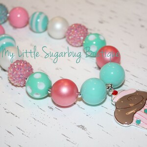 May include: A colorful necklace made of pink, white, and turquoise beads with a cupcake charm. The necklace is made of large, chunky beads and is perfect for a little girl.