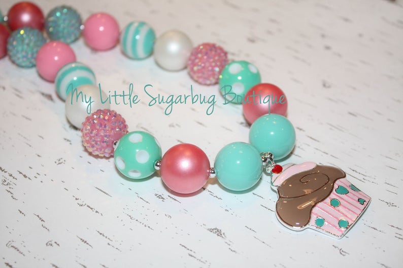 May include: A colorful necklace made of pink, white, and turquoise beads with a cupcake charm. The necklace is made of large, chunky beads and is perfect for a little girl.