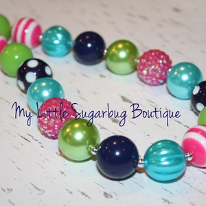 Pode incluir: Um colar colorido com contas grandes e redondas em várias cores e padrões. As contas incluem verde liso, azul marinho, verde-azulado e rosa com listras brancas ou bolinhas. Algumas contas são cobertas com lantejoulas rosa. O texto "My Little Sugarbug Boutique" é visível.