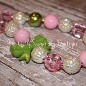 Puede incluir: Un collar de cuentas gruesas de color rosa, verde y blanco con un lazo verde. El collar está hecho de cuentas grandes y redondas y tiene una cadena plateada.