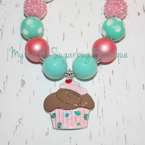 May include: A turquoise and pink beaded necklace with a cupcake charm. The cupcake is brown with pink frosting and white sprinkles. The necklace is made of large, round beads.
