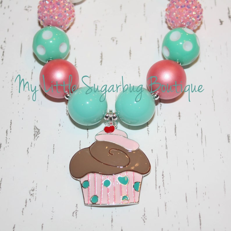 May include: A turquoise and pink beaded necklace with a cupcake charm. The cupcake is brown with pink frosting and white sprinkles. The necklace is made of large, round beads.