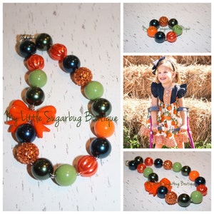 Puede incluir: Un collar colorido con cuentas verdes, naranjas y negras. El collar tiene un gran lazo naranja y una cuenta brillante. El collar se llama "My Little Sugarbug Boutique".
