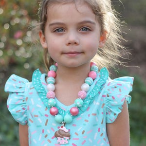 May include: A young girl wearing a light blue dress with a pink and white cupcake print and a pink headband. She is wearing a multi-colored beaded necklace with a cupcake charm.
