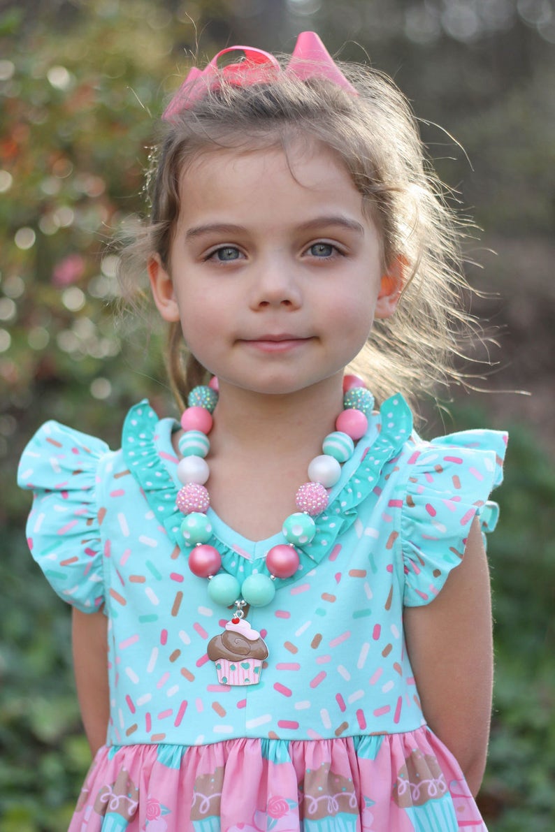 May include: A young girl wearing a light blue dress with a pink and white cupcake print and a pink headband. She is wearing a multi-colored beaded necklace with a cupcake charm.