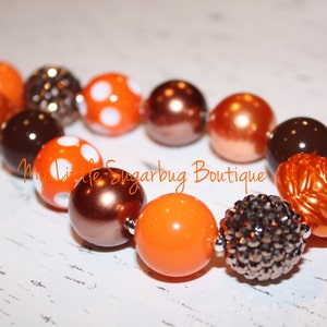 May include: A necklace with large, spherical beads in orange, brown, and silver. Some beads are plain, others have white spots, and some are textured. The necklace is displayed on a white surface. The text "My Little Sugarbug Boutique" is visible.