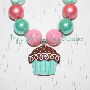 May include: A pink and teal beaded necklace with a cupcake charm. The cupcake is brown with white frosting and sprinkles.