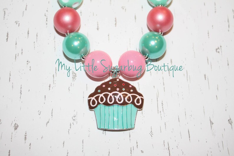 May include: A pink and teal beaded necklace with a cupcake charm. The cupcake is brown with white frosting and sprinkles.