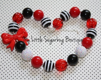 May include: A red and black chunky bead necklace with a red bow charm. The necklace is made with large, round beads in a variety of textures, including smooth, textured, and sparkly. The beads are alternating in a pattern of red, black, and white striped beads.