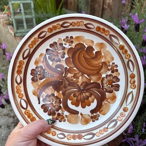 Könnte beinhalten: Ein weißer Keramikteller mit braunem und orangefarbenem Blumendesign. Der Teller hat einen gewellten Rand und einen braunen Rand.