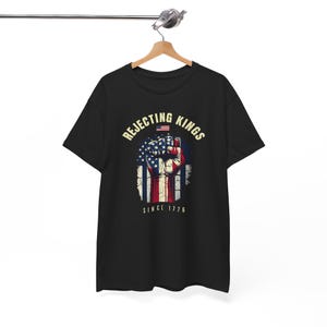 May include: A black short-sleeved t-shirt featuring a graphic of a raised fist coloured with the American flag pattern. The text REJECTING KINGS is arched above the fist, with a small flag icon and the text SINCE 1776 printed below the fist.