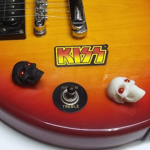 Peut inclure: Un autocollant de logo KISS rouge, blanc et noir sur une guitare électrique sunburst avec un bouton de crâne noir aux yeux rouges et un bouton de crâne blanc aux yeux rouges. La guitare a un micro noir et un sélecteur à bascule marqué "Rhythm" et "Treble".