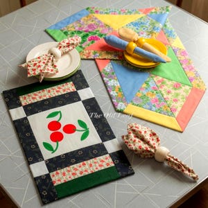 May include: A rectangular placemat with a central cherry graphic, surrounded by black floral and green borders. Includes a colorful geometric table runner, two white plates with floral napkins in wooden rings, and two yellow plates with blue napkins.