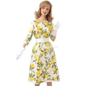May include: A cream-colored midi dress featuring a vibrant yellow lemon and green leaf print, three-quarter length sleeves, a boat neckline, and a wide white belt at the waist. The garment is paired with white wrist-length gloves.