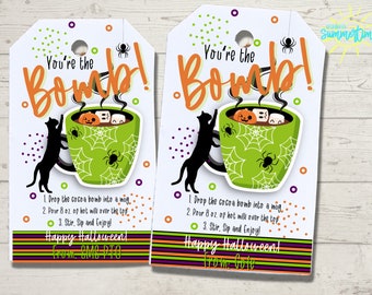 May include: Two printable gift tags with a green mug with a spiderweb design and a black cat reaching for the mug. The mug has a steaming cup of hot chocolate with marshmallows and a jack-o-lantern inside. The text on the tags reads "You're the Bomb!" and includes instructions for making hot chocolate. The tags are decorated with orange, purple, and green stripes and polka dots. Happy Halloween! From: SMC PTO
